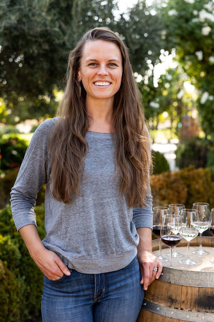 With a friendly smile, Laura leans on a wine barrel.