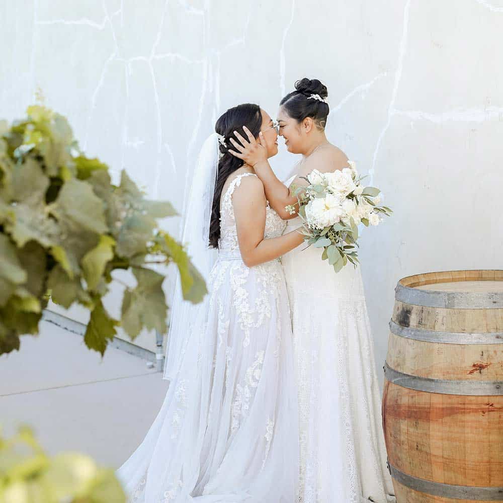 The brides both in white dresses look into each others eye and smile as they embrace at their outdoor winery wedding in the Sacramento Delta.