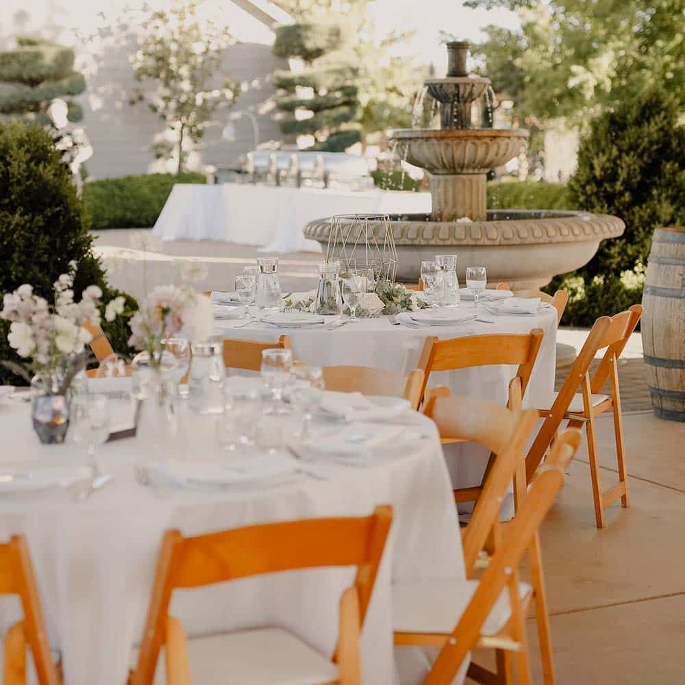 Tables set in white linens with bud vases and small flowers in them with a three-tiered fountain in the background at this outdoor winery wedding.