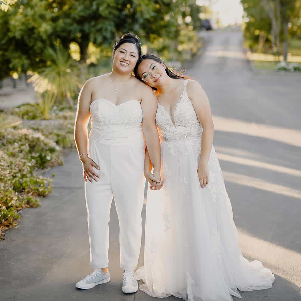 The bride and bride holds hands and one of them puts her head on the others shoulders as they stand at the bottom of a driveway lined with mature trees at their outdoor winery wedding.
