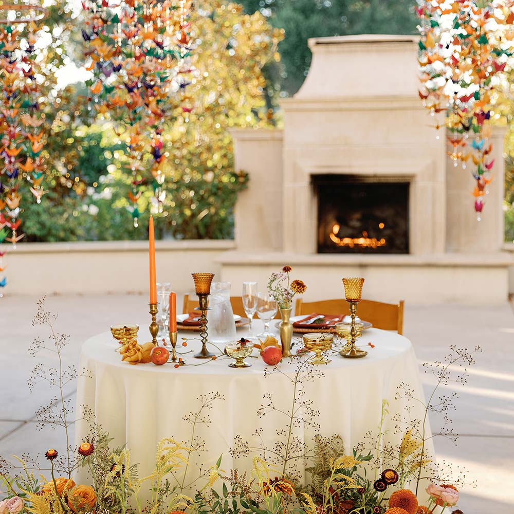 The head table with the lit outdoor fireplace in the background are surrounded by orange and green flowers to the front of their table with their table set in orange glassware and candles at their outdoor Sacramento winery wedding.