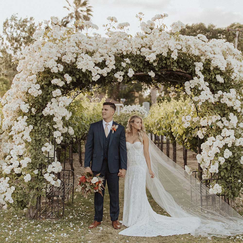 A bride leans into a groom as he holds her bouquet as they stand under an arch in full bloom of white carpet roses.