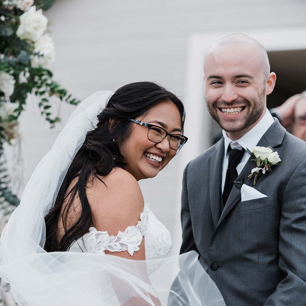 The bride and groom both smile into the camera as her veil flies in the wind.