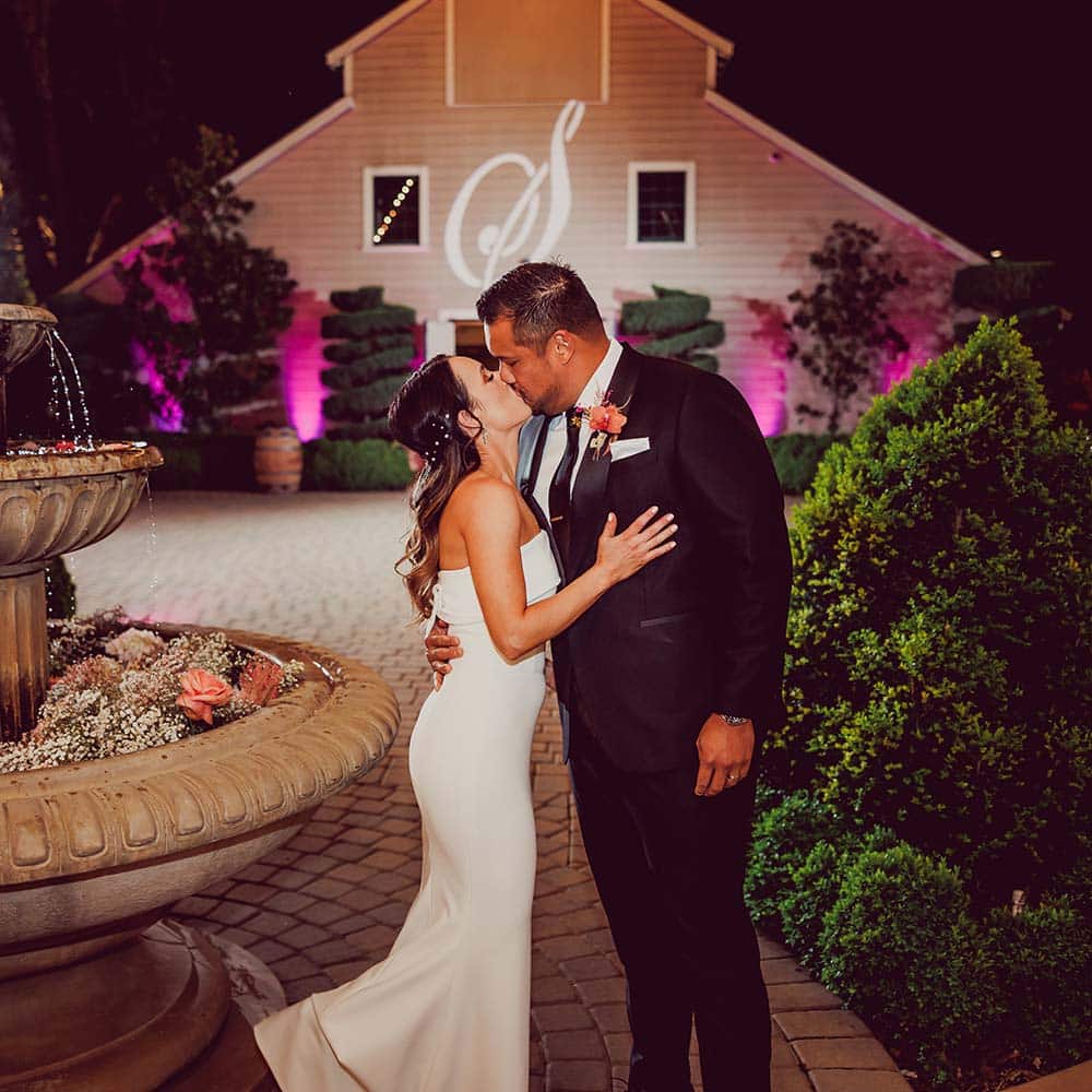 The brides and groom share a kiss in front of a 1918 bar with the initial lit up on the bar as they stand next to a three tiered fountain which is full of baby's breath and pink roses.