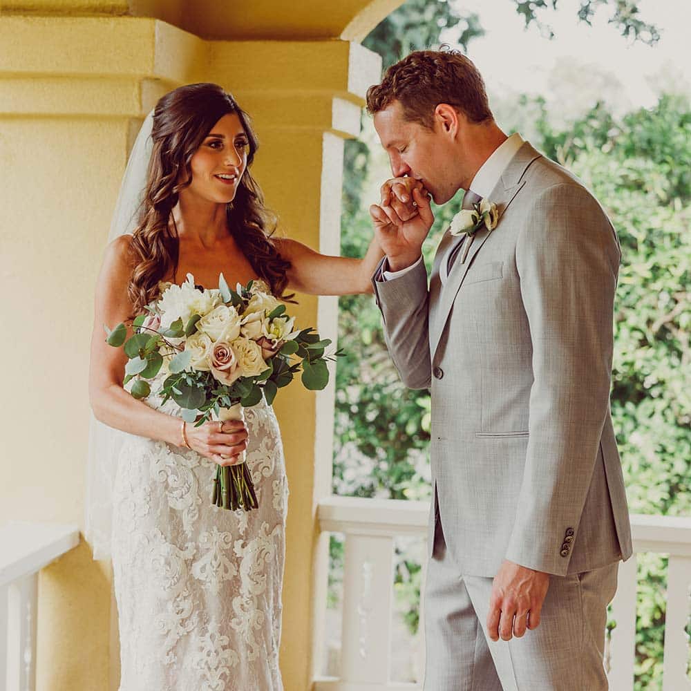 A groom and his bride on the porch of a 1920's craftsman home as she looks at him as he kisses her hand.