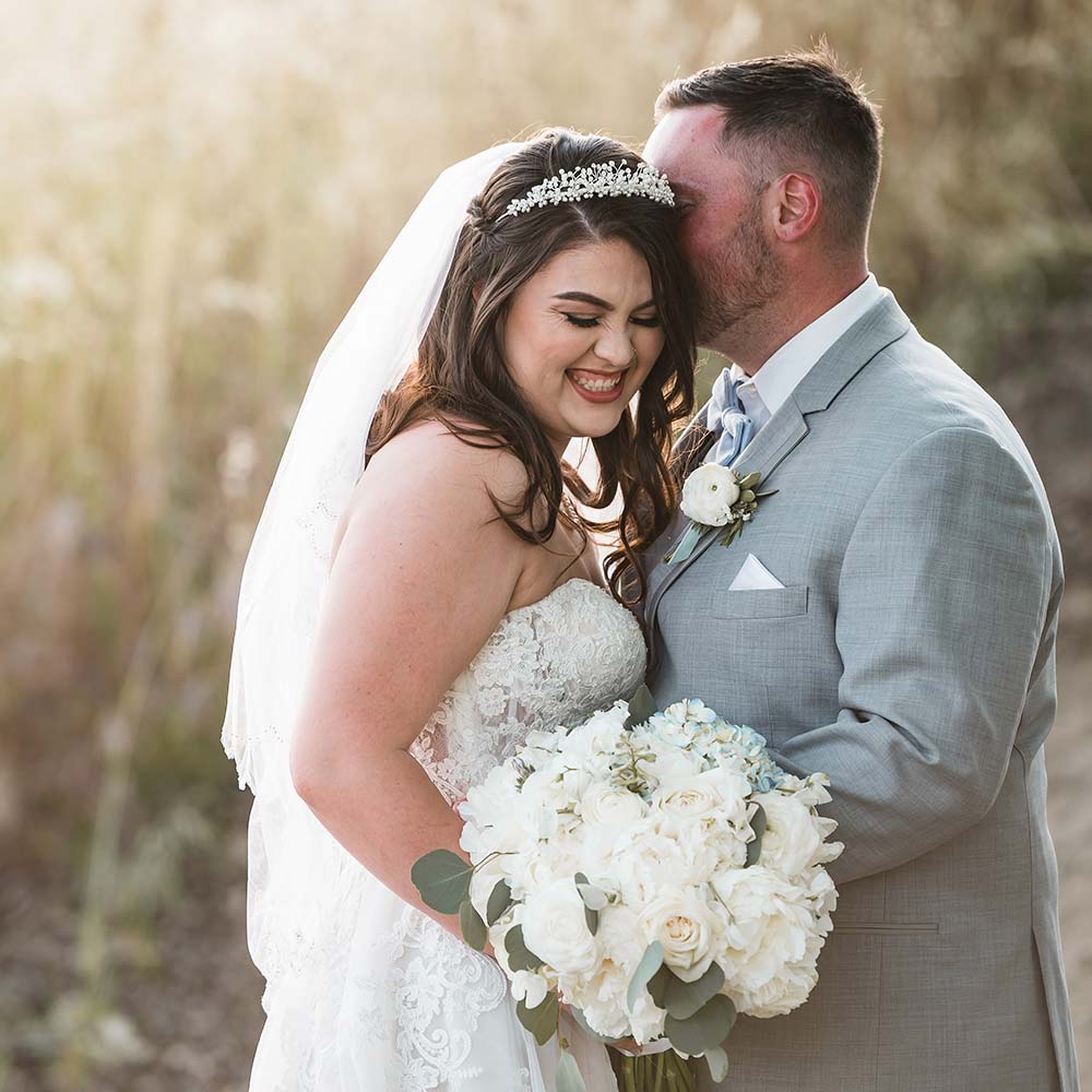 A groom holds his bride so you cannot see his face but his bride has a full small as their backdrop is a wheat field.