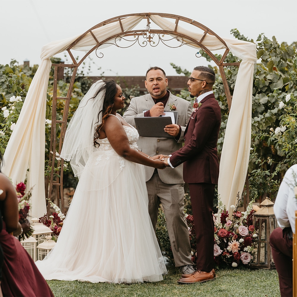 The bride and groom exchanging their vows in front of the officiant and guests and under the pergola at the winery wedding in Sacramento.