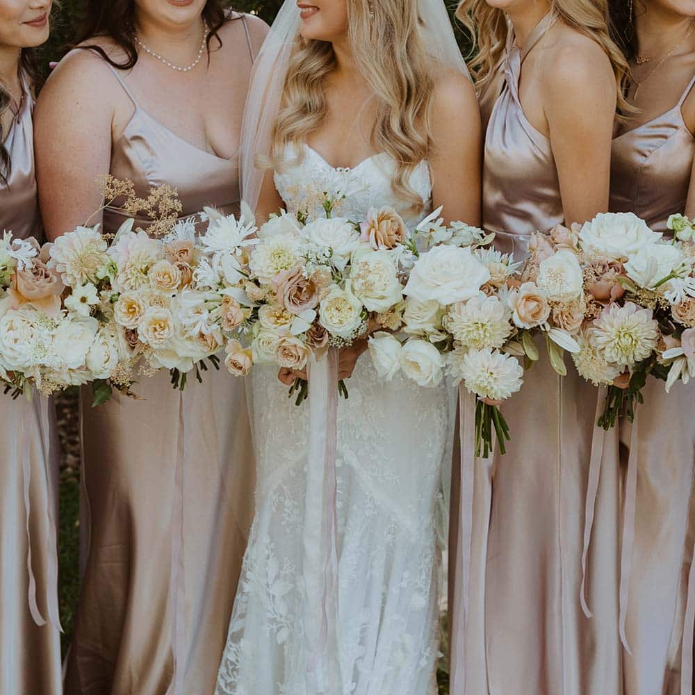 The brides centered by her 6 bridesmaids in beige dresses and holding full bouquets with white, beige and soft pink flowers at their outdoor winery wedding in Sacramento.