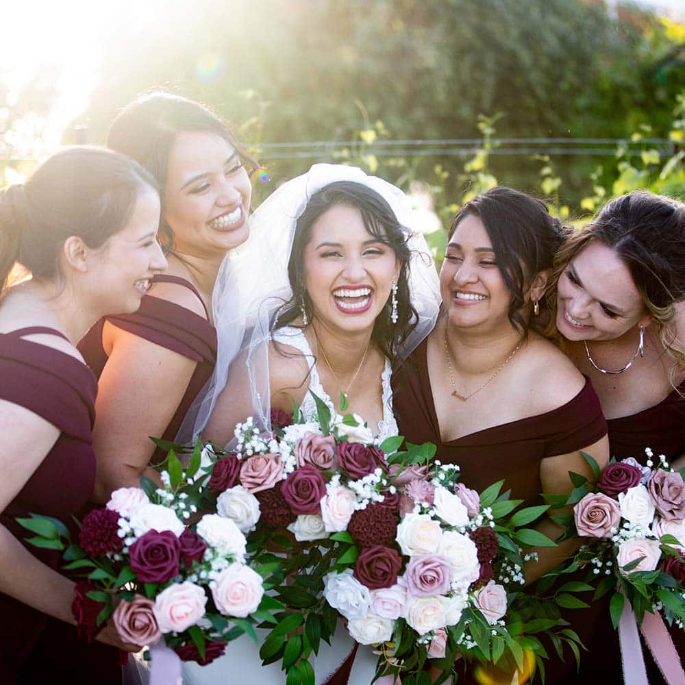 The bride smiling as she is surrounded by her bridesmaids in a sea of white and burgundy bouquets.