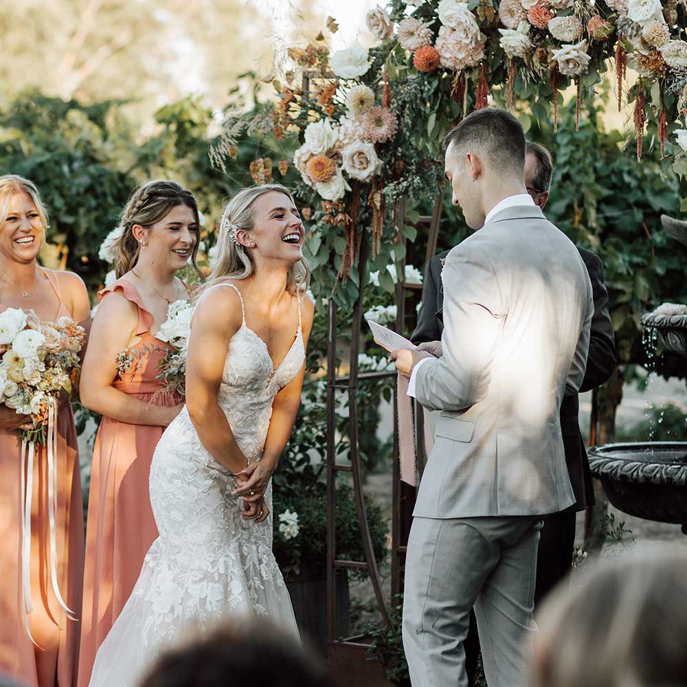 As the groom says his vows, the bride bends over with a big smile and her bridesmaids join her in smiling.