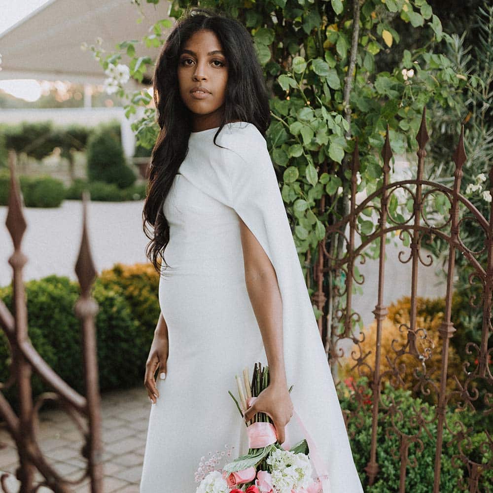 A bride in her long white cape dress holding her pink and white full bouquet as she stands at the wrought iron gates with the covered tent as a backdrop.