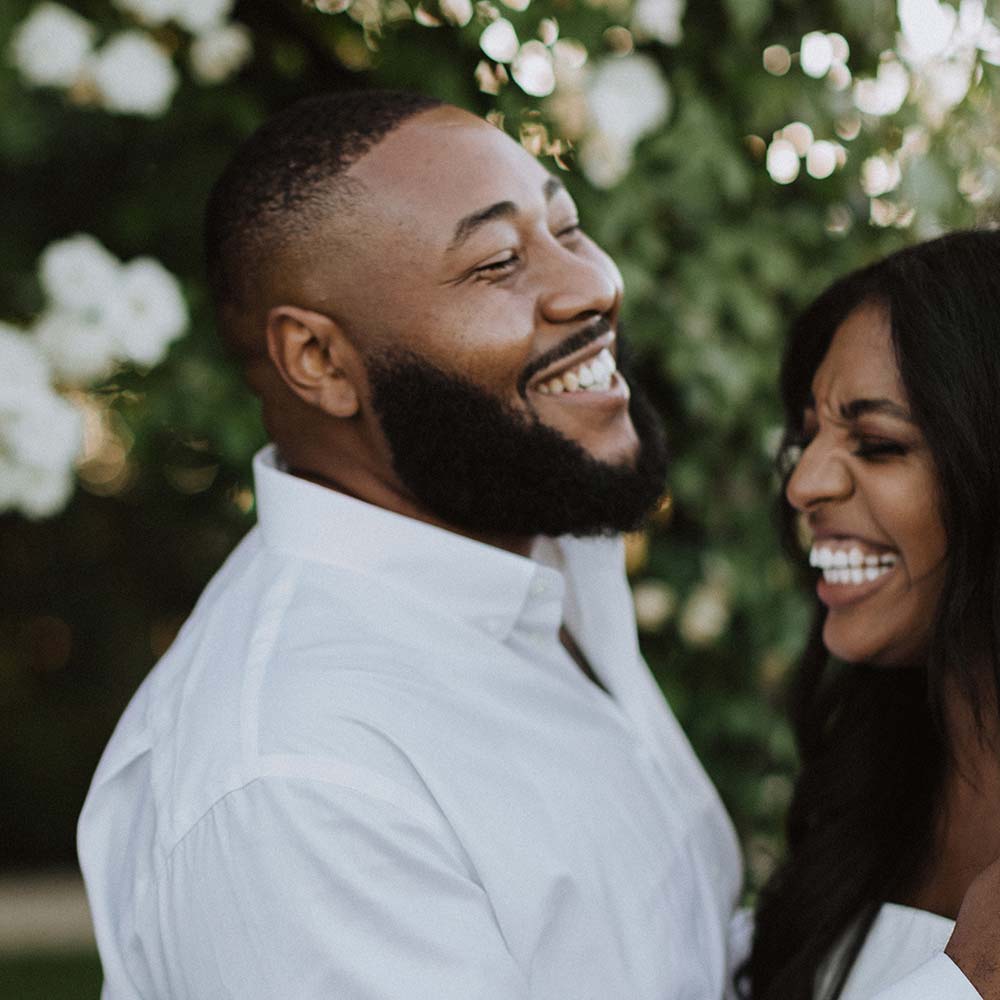 A groom holds his wife as they share a warm smile under the while carpet rose arches at this outdoor wedding venue.