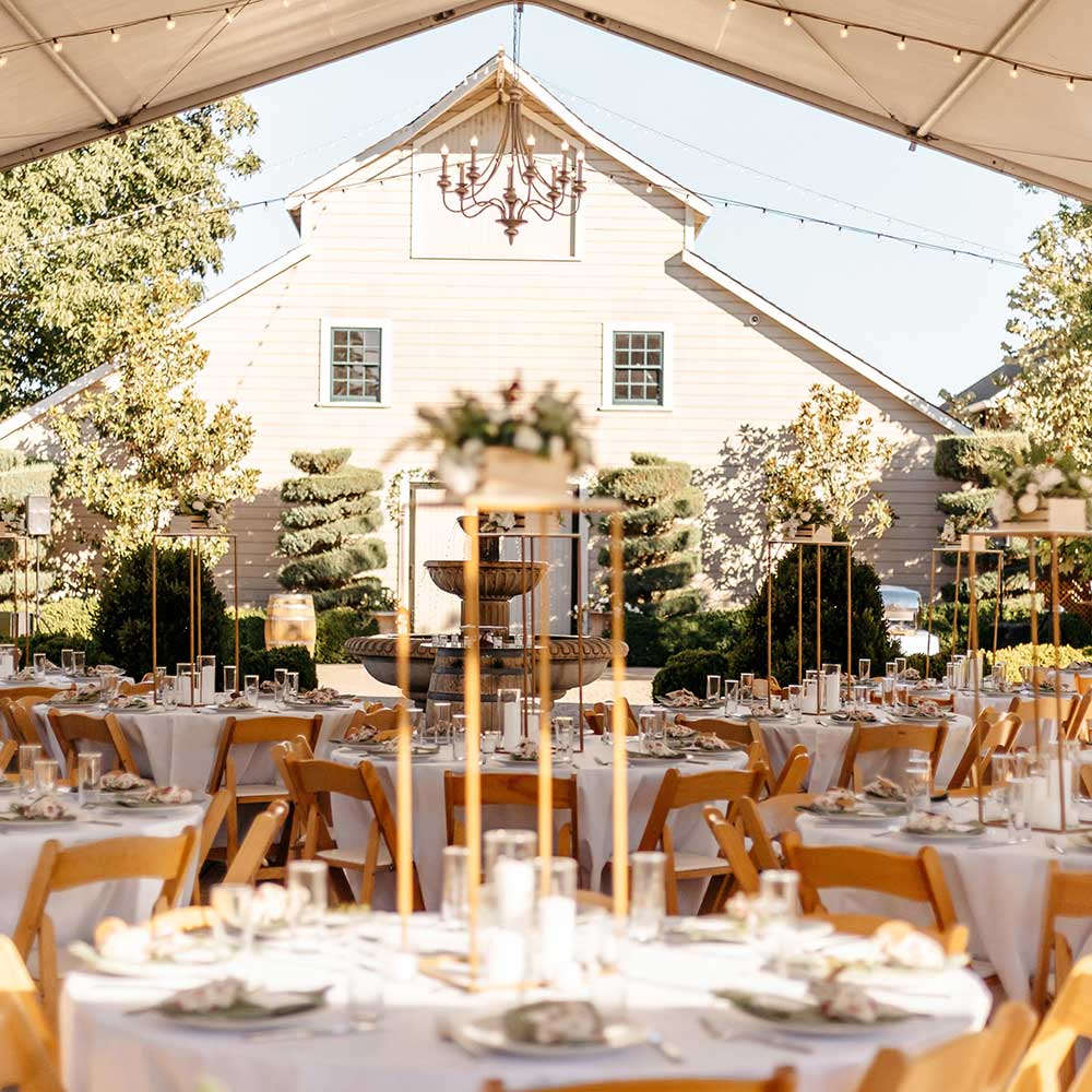 A full views of the wedding guest tables looking onto the 1920's winery with the tables under an tent with market lights at this outdoor winery wedding.