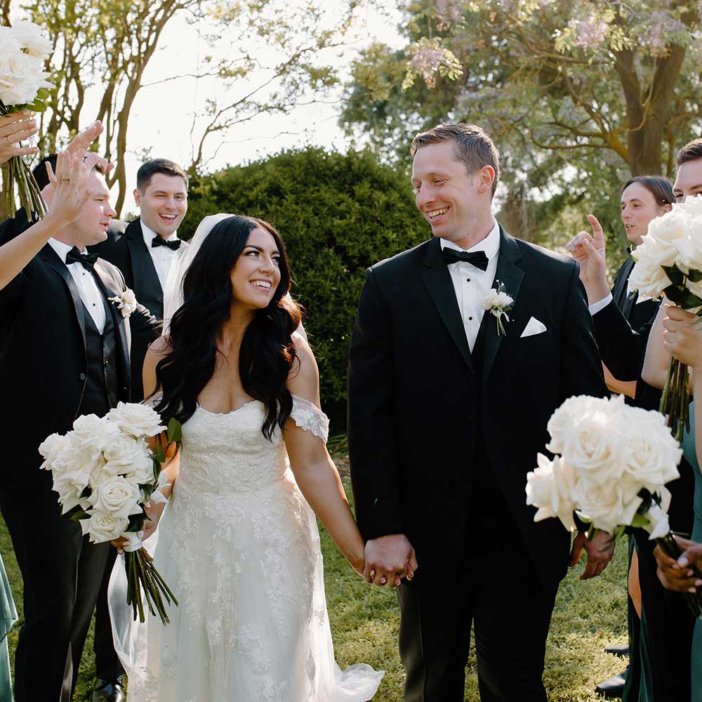 The bride and groom hold hands and smile while their wedding parties smile an cheer them on.