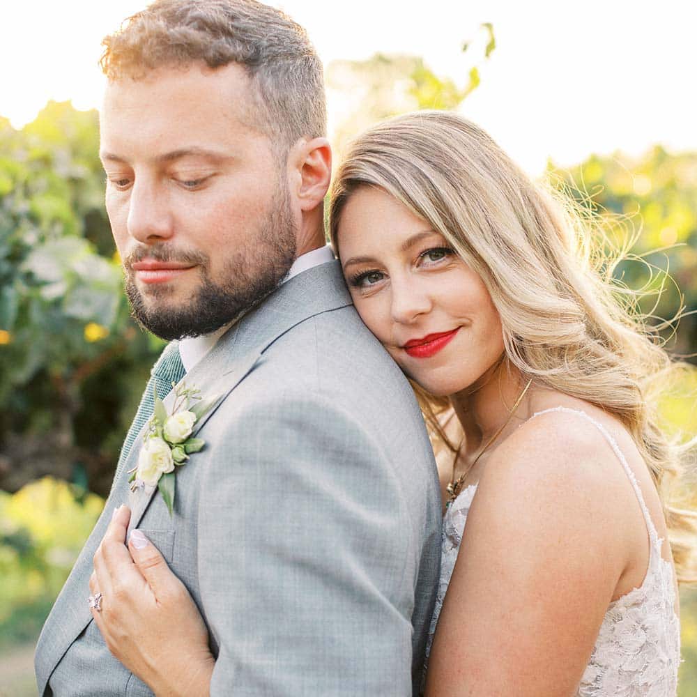 A close up of a bride holding a groom from the back as she smiles into the camera and he look down.