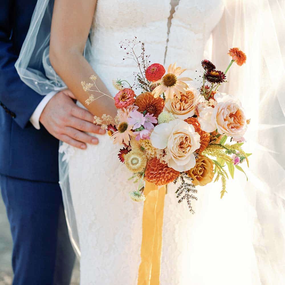 The groom with his hand on his new bride's waist took forward as she holds her bouquet full of orange, pink, purple and yellow flowers at the winery wedding in Sacramento.