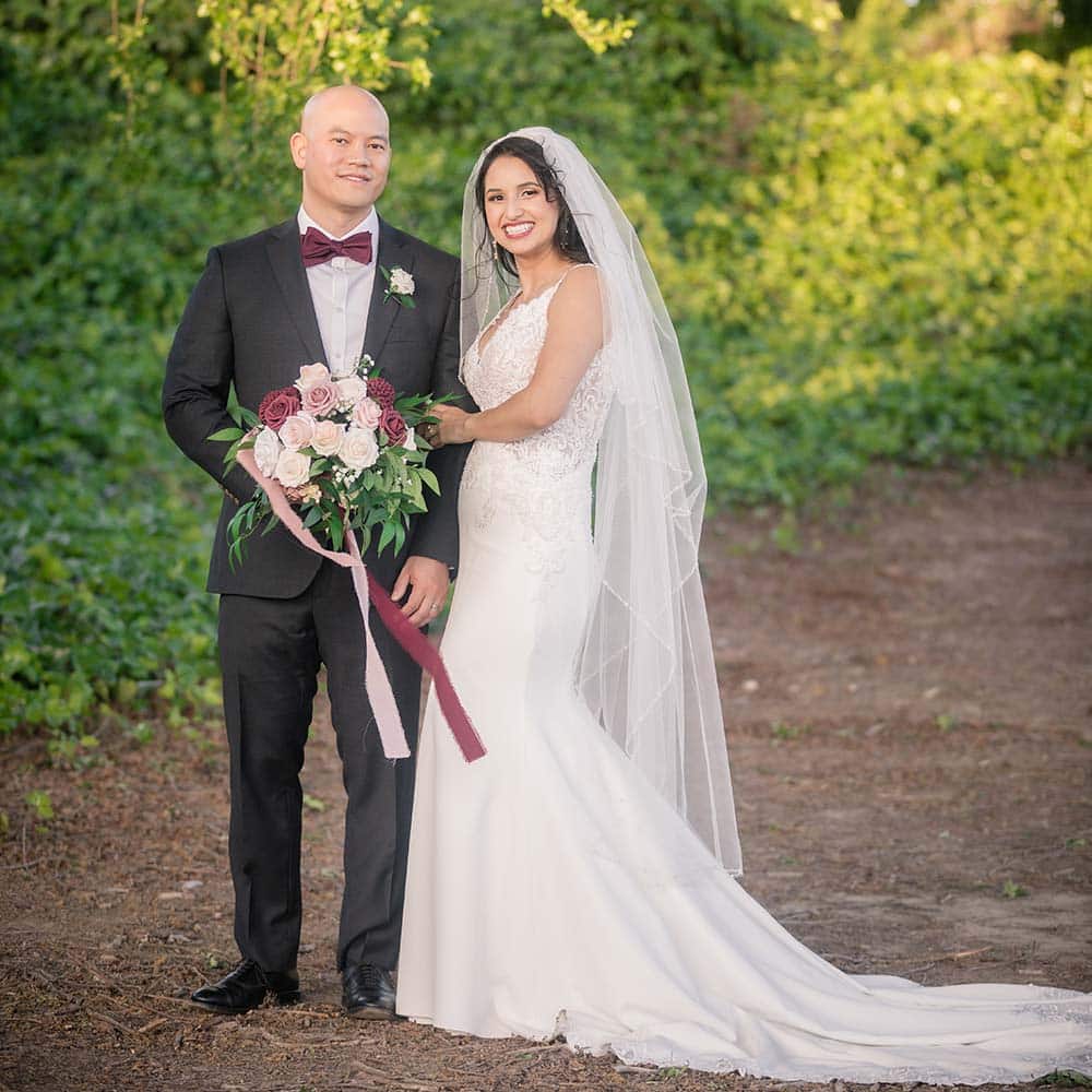 The bride holds onto the grooms arm as he holds her bouquet and they look at the camera with big smiles on their faces.