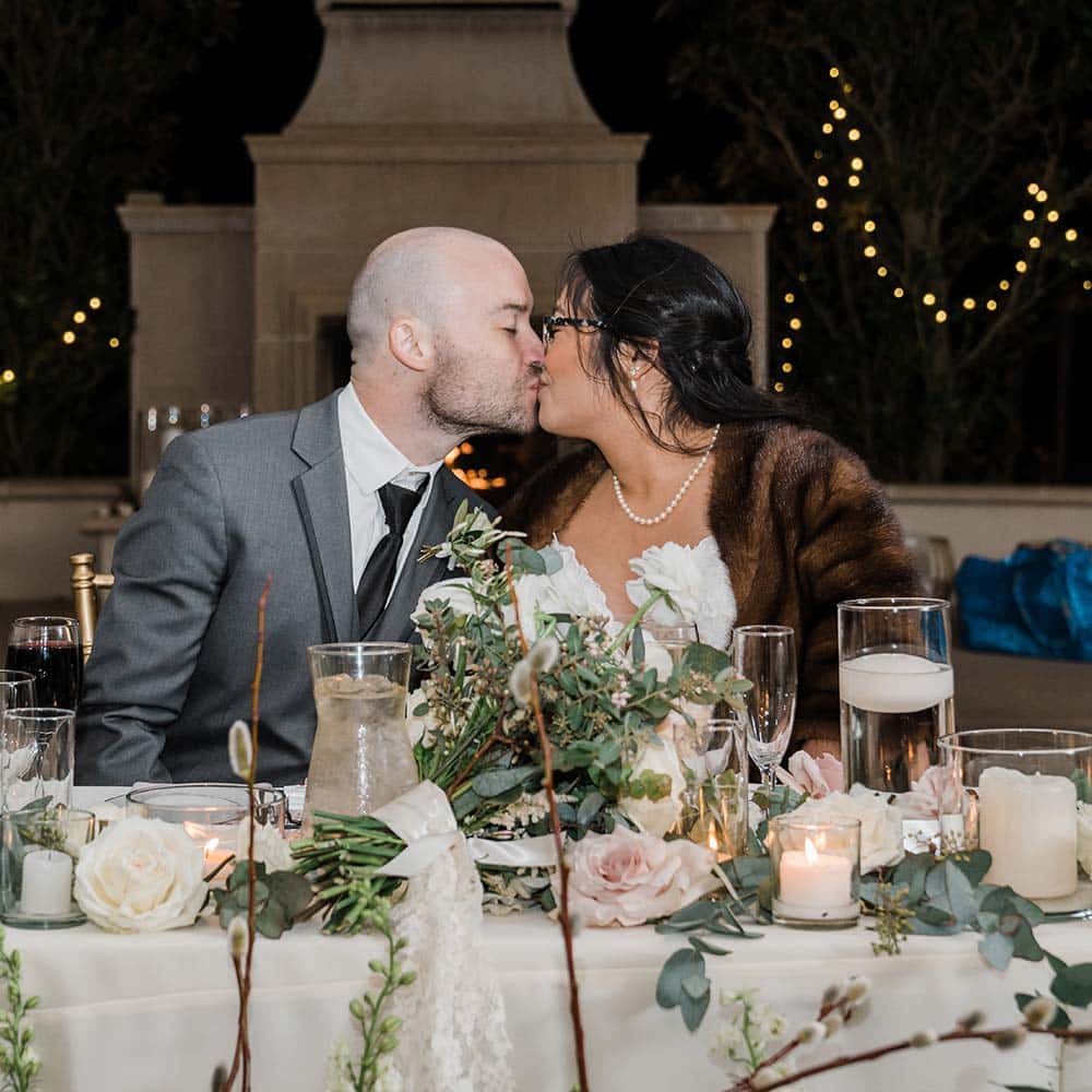 The couple kiss at their head table which is adorned with flowers and crystal glass and votive vases and market lights and the lit fireplace are seen in the background.
