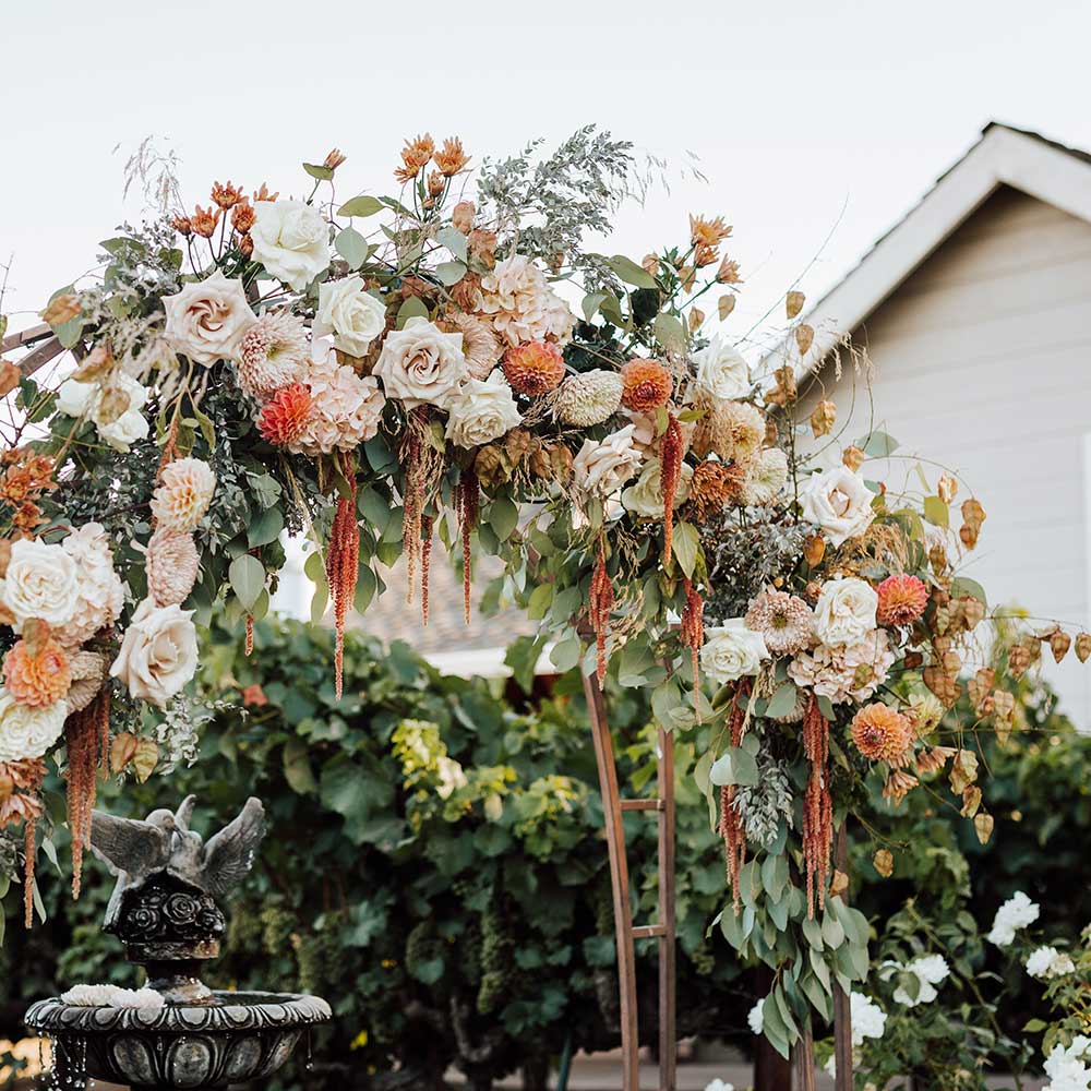 A wedding arch fully covered in orange and beige flowers with greenery and a small fountain with doves on top can be seen at the bottom of the arch.