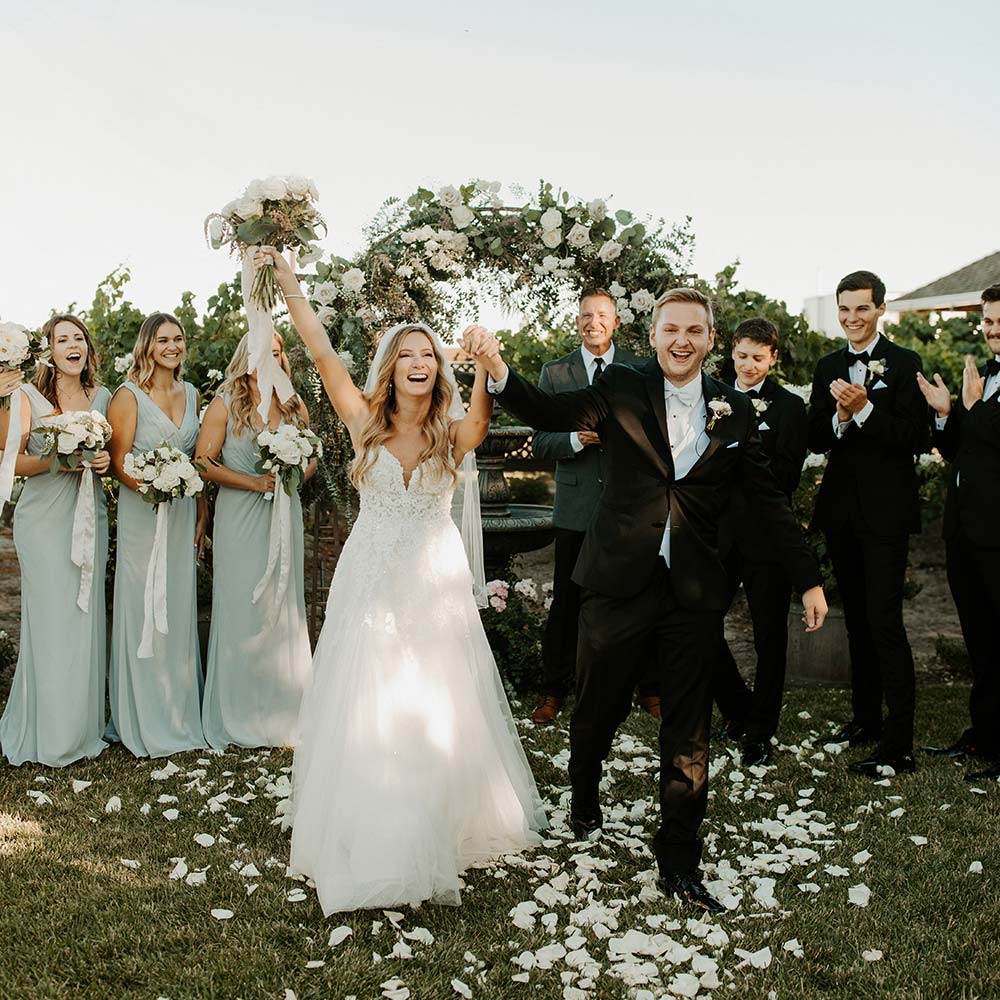 A bride and from smiling with their arms in the air after they say their vows. The flowered arch in is the background as are their groomsmen and bridesmaids.