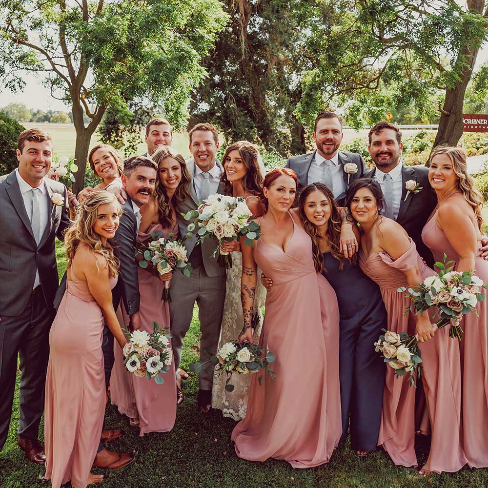 The wedding party and bride and groom all gather together on the green lawn with mature trees in the background.