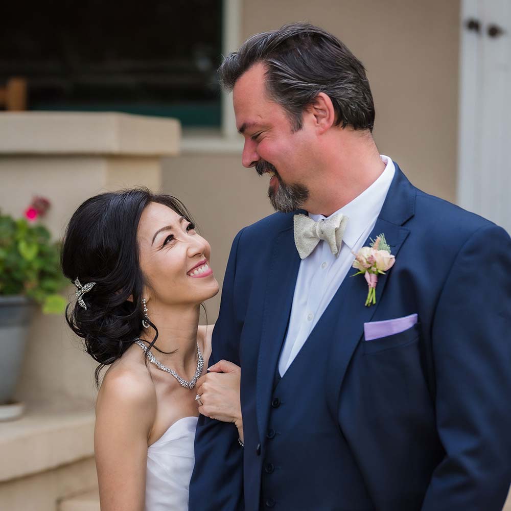 A bride holds onto her husbands arms and looks up at him smiling as they enjoy a bit of privacy before seeing their guests at their outdoor winery wedding.