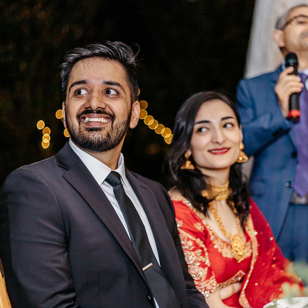 The evening shows the market lights in the background as the bride and groom smile while their parents in the background give a speech about them.