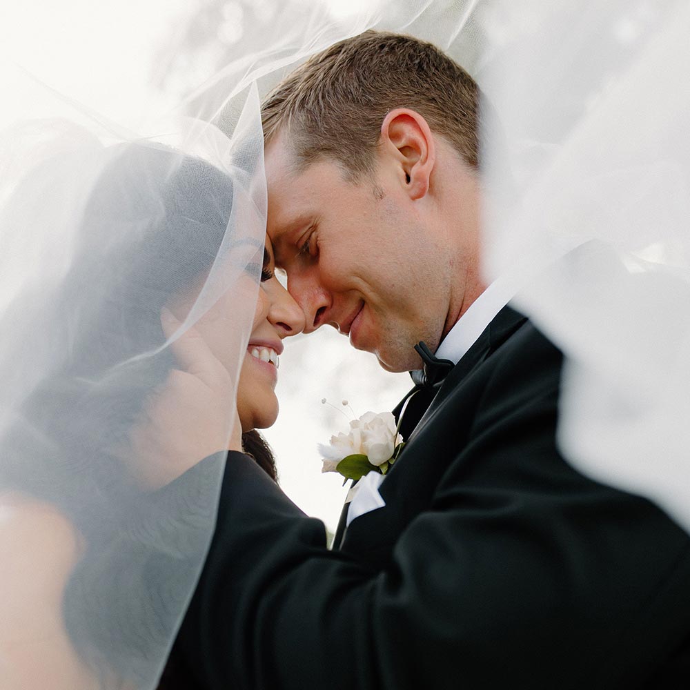 A groom holds his brides neck with his hand and they embrace under her veil.