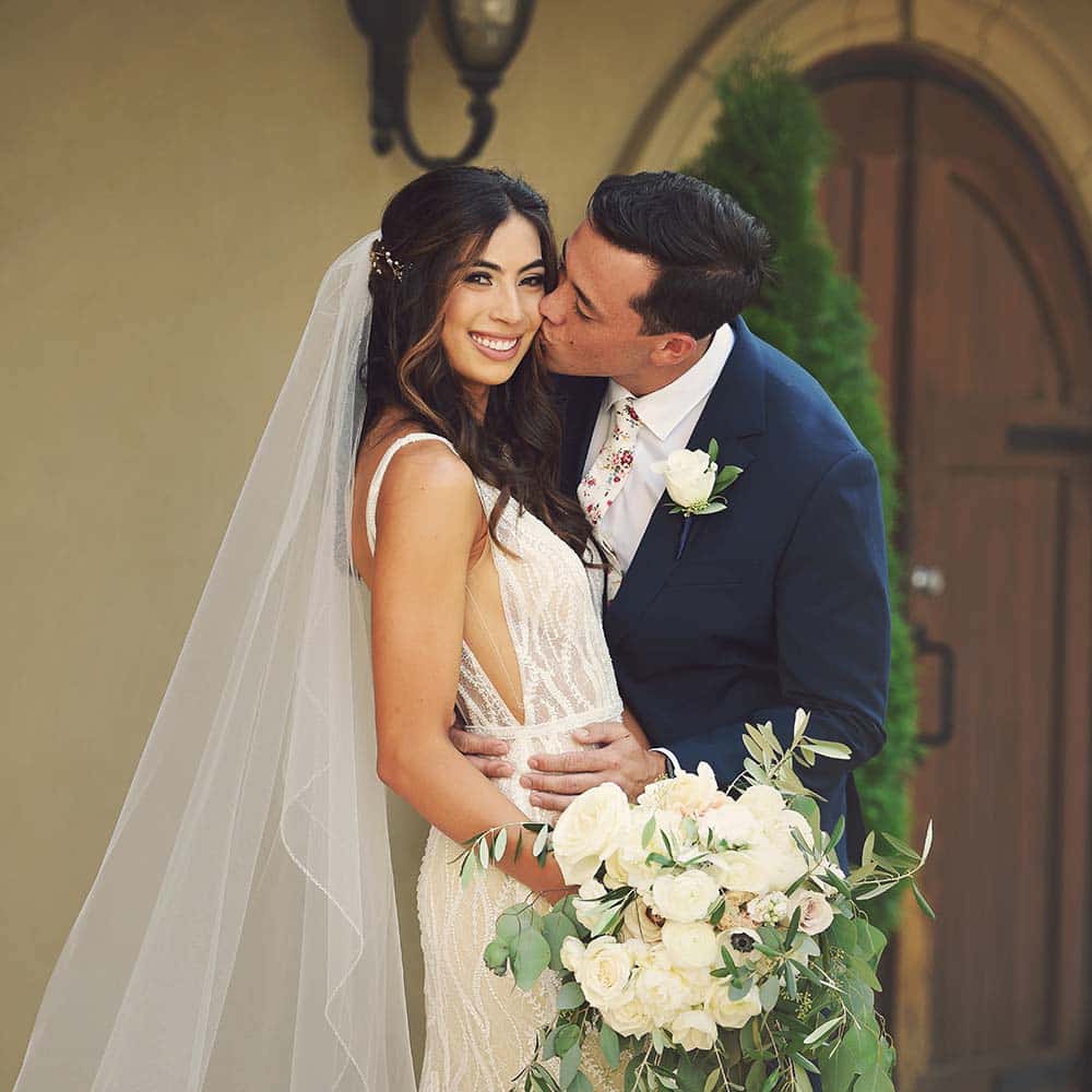 A groom holds his wife around her waist and as he kisses her, she smiles into the camera.