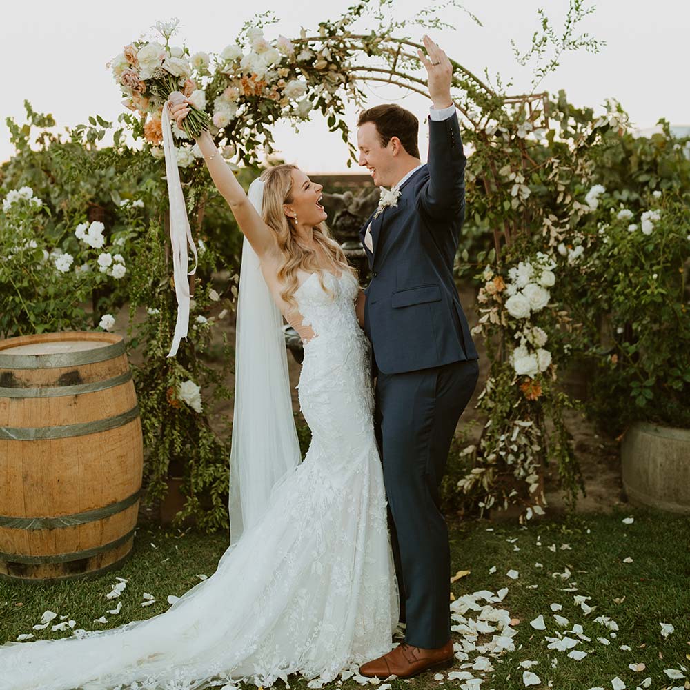 The bride and groom raise their hands in the air just after they exchange vows with her bouquet up in the air and their floral arch in the background.