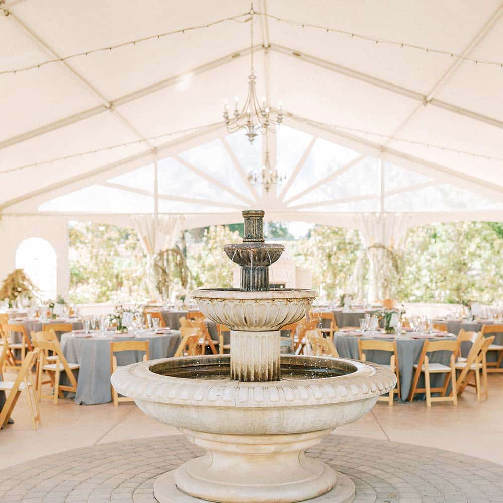 Looking toward a three tier outdoor fountain are many round tables set up under a covered tent.