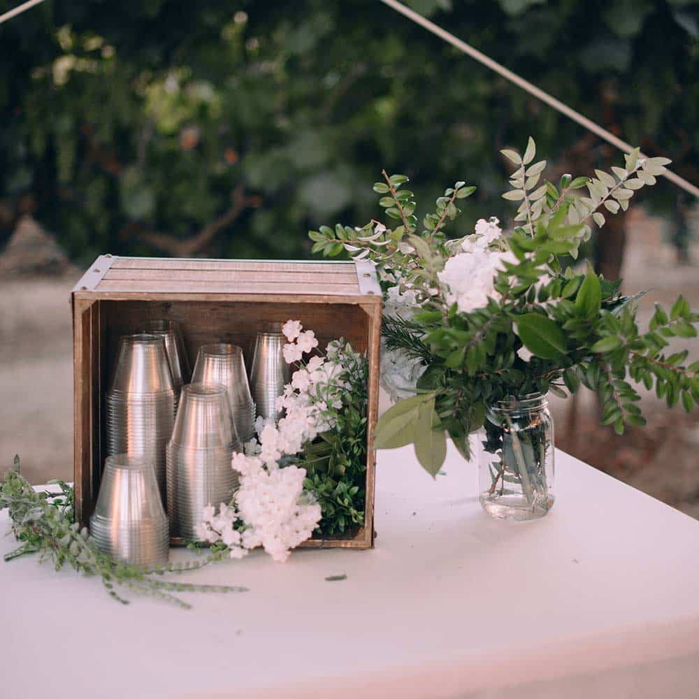 A wooden crate with plastic cups and white flowers and greenery sits on a white linen table cloth.