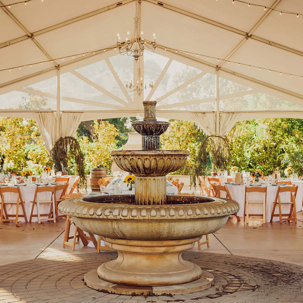 A three tiered fountain with water coming out of it sits in front of white linen round tables which are decorated with yellow daisies.