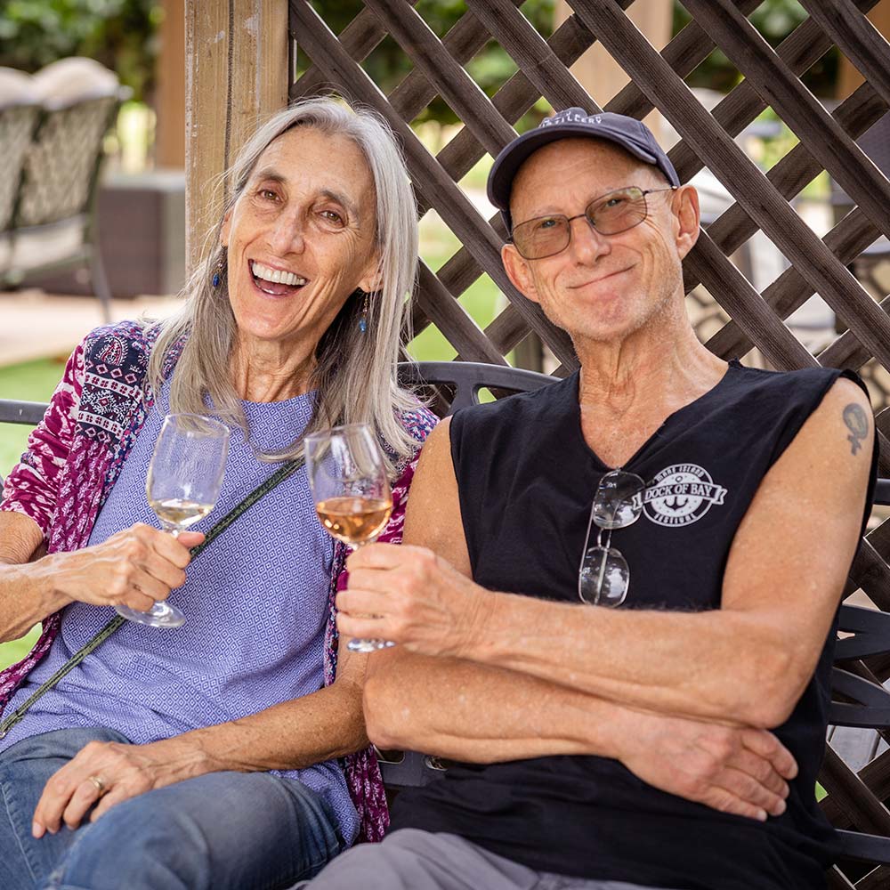A couple leans into each other and smiles into the camera and they toast with their glasses of white wine.