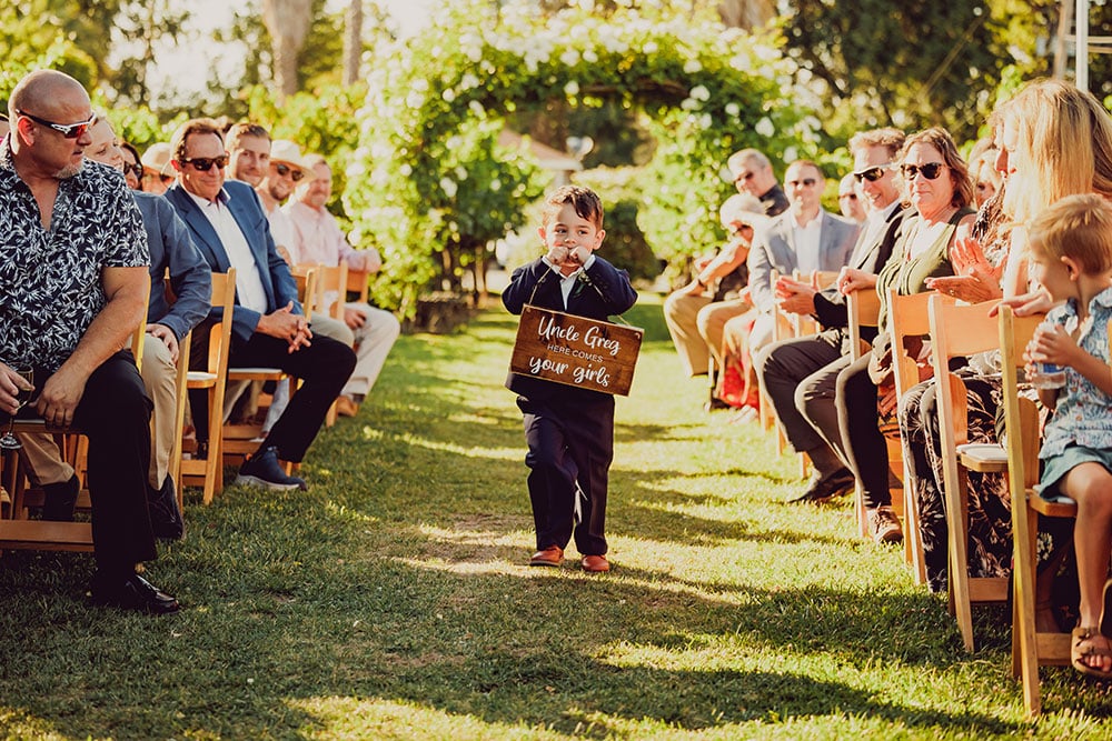 Shy ringbearer walks down the vineyard wedding aisle as wedding guests smile.