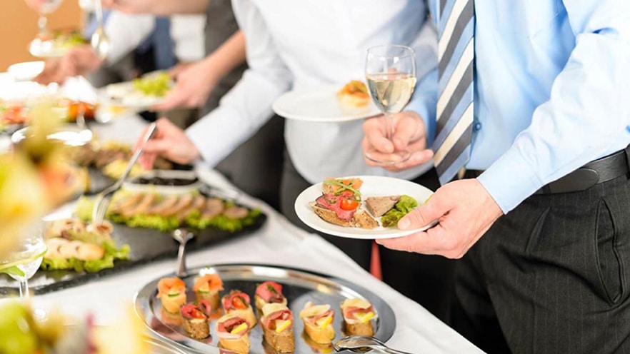 A man is holding a small plate of food in one hand and a glass of white wine in the other hand as he goes through a buffet line at a corporate event.