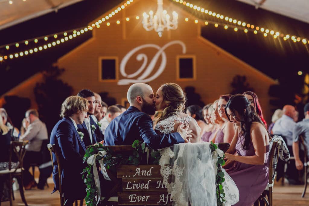 Bride and Groom kiss at table during outdoor tent wedding reception with string lights.