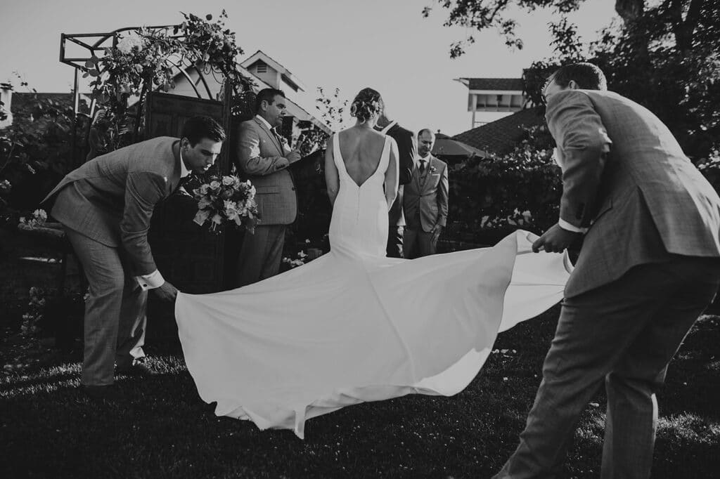 Two male attendants in gray suits fluff out the bride's train at an outdoor wedding ceremony site in the Sacramento Delta.