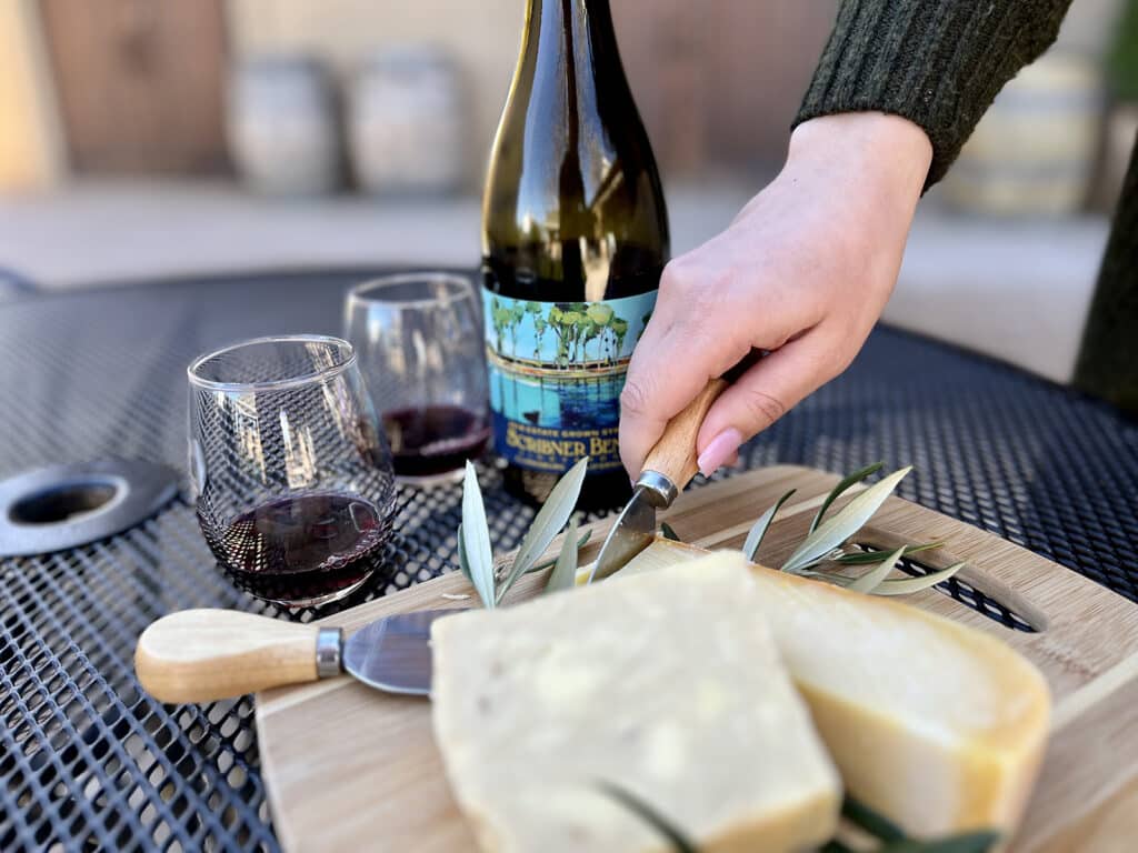 A woman's hand cuts cheese on a board in front of a two glasses and a bottle of Scribner Bend Vineyards Syrah.