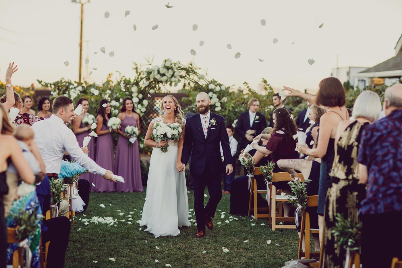Smiling bride and groom walk down the wedding aisle at Scribner Bend Vineyards as guests toss rose petals in the air.