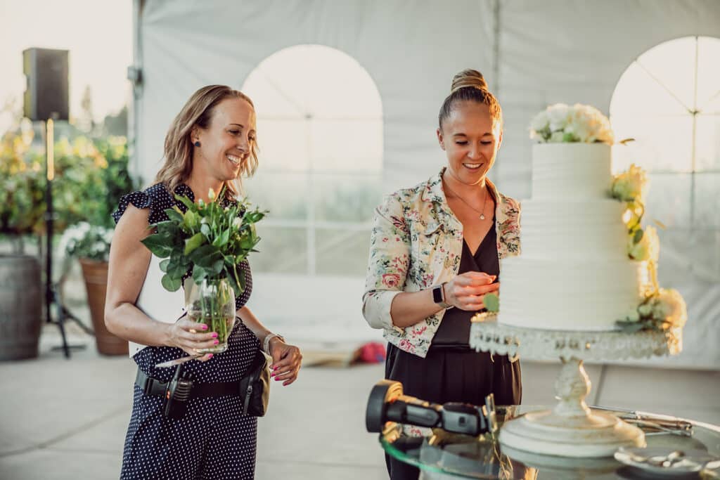 Two female wedding staff at Scribner Bend Vineyards assist with setting up the cake table at an outdoor wedding.
