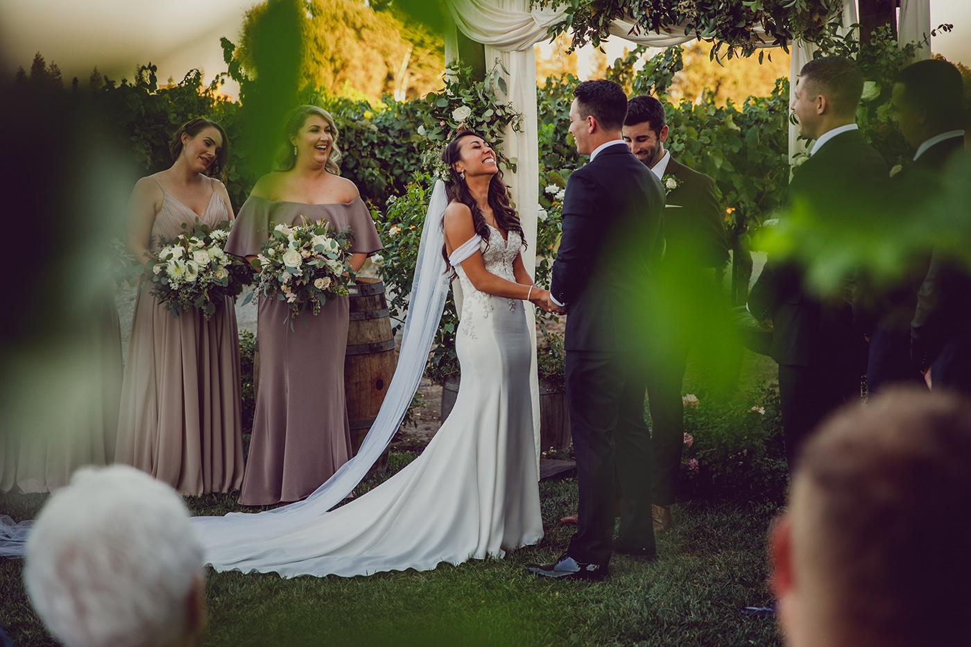 Bride and groom are laughing at the altar of their outdoor wedding, with grape leaves in the foreground.
