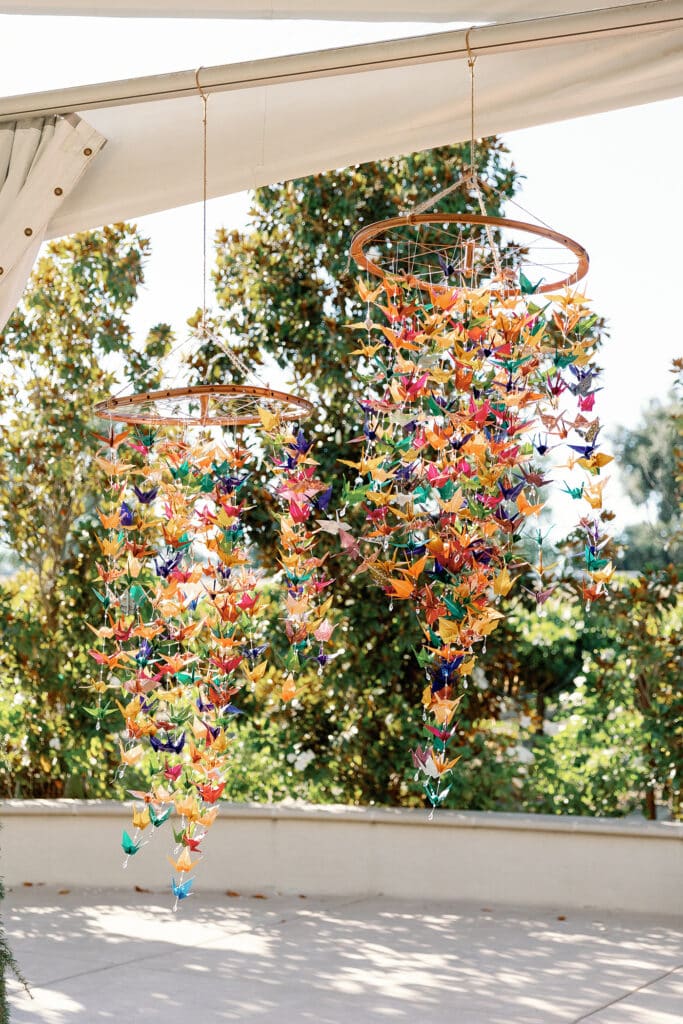Hundreds of colorful paper cranes hang from bicycle wheels at an outdoor wedding reception venue.