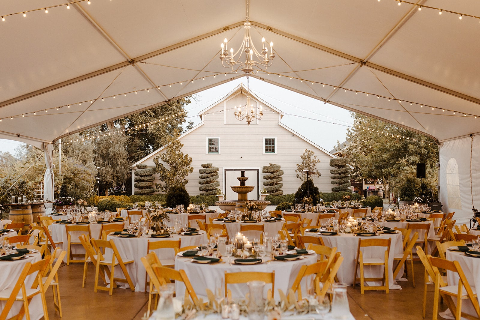 An outdoor wedding reception is set up under a tent strung with lights and a barn in the background.